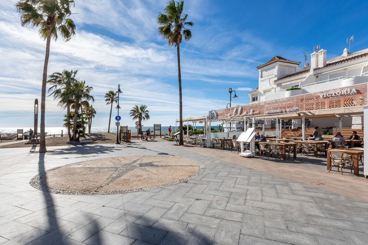 Un paseo marítimo en la playa con palmeras, mesas al aire libre y un edificio con un cartel que dice 'VICTORIA'.