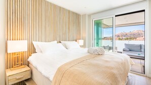 A hotel room with a large bed, a nightstand with a lamp, a view of the city through the window, and a balcony with a sofa.