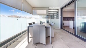 A modern dining area with a large table and chairs, a view of the sea, and a glass door leading to a kitchen.