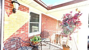 A covered porch with two metal chairs, a potted plant, and a brick wall. - Condo Comforts of HOMME (Kannapolis)