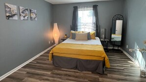 A bedroom with a bed, a bedside table, a mirror, and a window with curtains. - Condo Comforts of HOMME (Kannapolis)