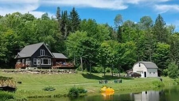 A house with a large deck and a smaller building nearby, surrounded by trees and a pond. - Le Rustique avec lac privé (Château-Richer)