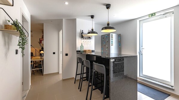 A modern kitchen with a bar area, featuring pendant lights, a refrigerator with magnets, and a door with an emergency exit sign. - Amazing 3BDR Duplex in Lisbon by LovelyStay (Lisbon)