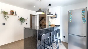 A modern kitchen with a central island, bar stools, pendant lighting, a refrigerator with magnets, and a staircase leading to another level.