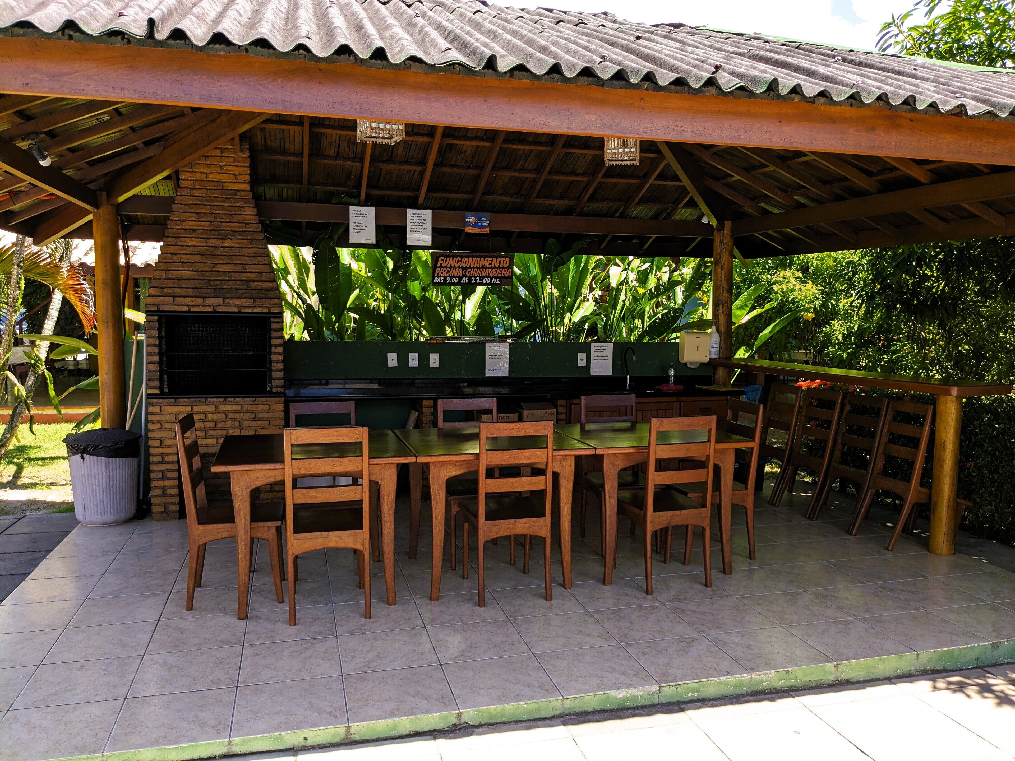 An outdoor dining area with wooden tables and chairs, a brick wall, and a green planter with tropical plants.