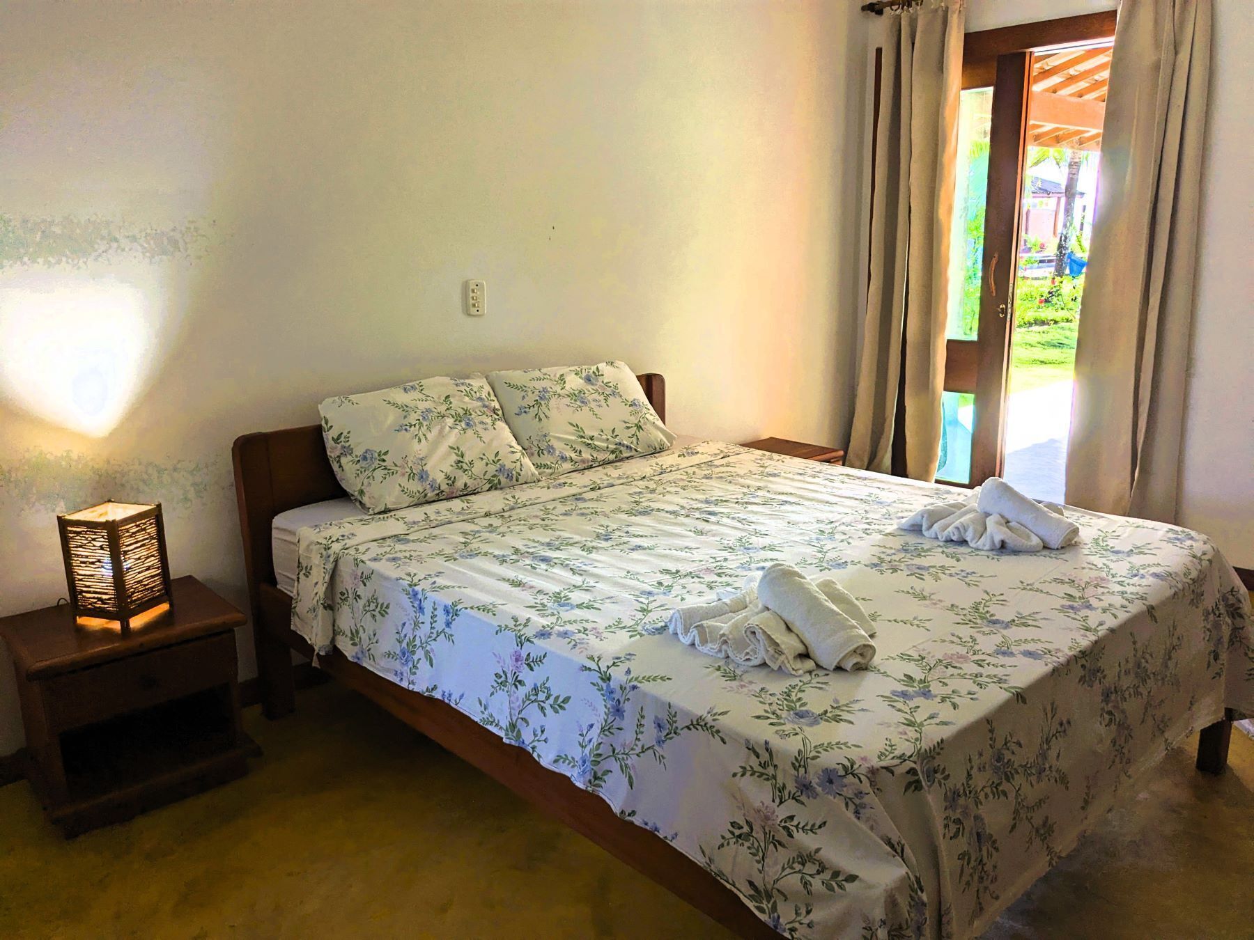 Quarto com cama, mesa de cabeceira, abajur e vista para o exterior.