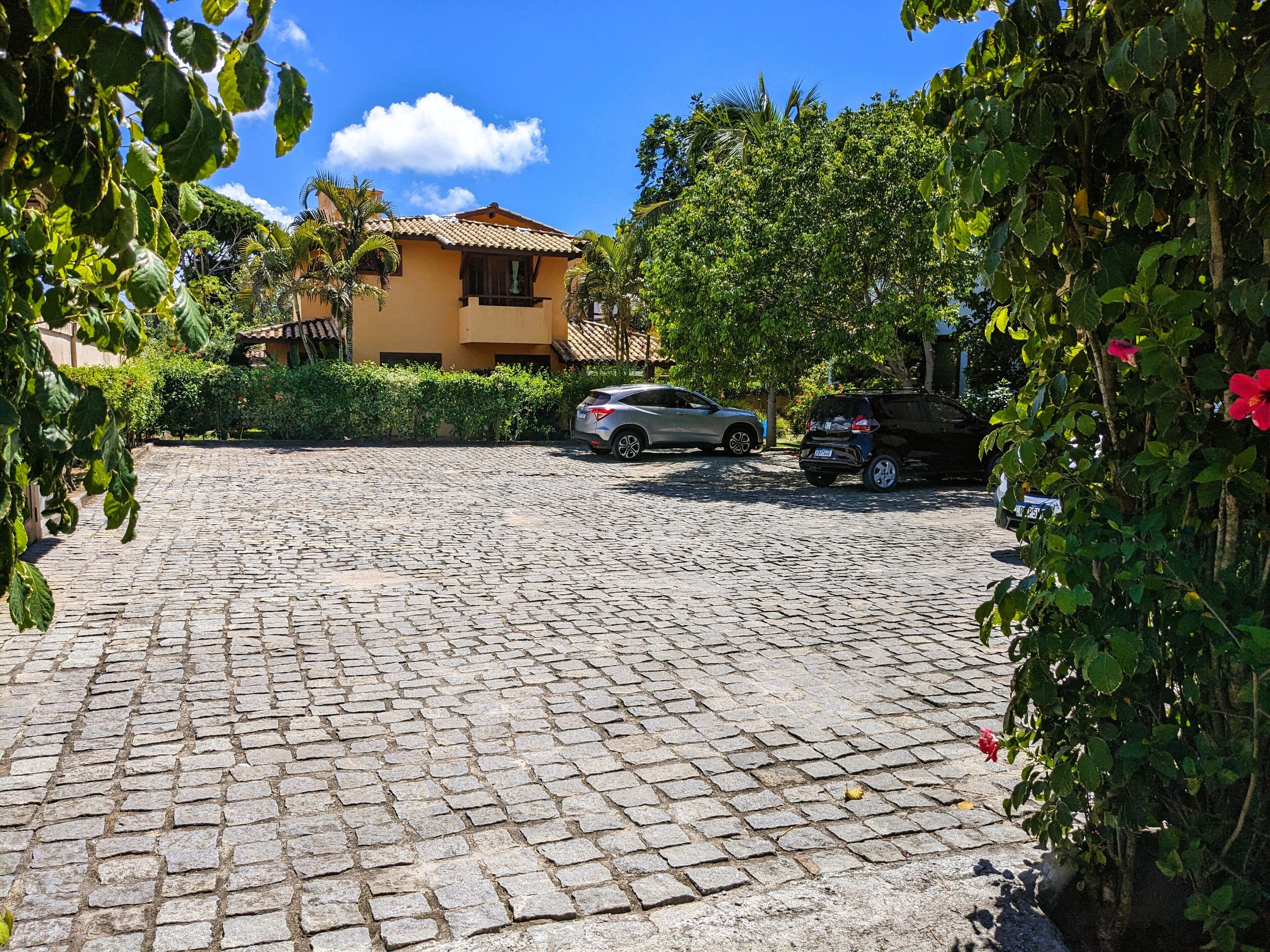 A cobblestone driveway leading to a yellow building with a tiled roof, surrounded by greenery and parked cars.