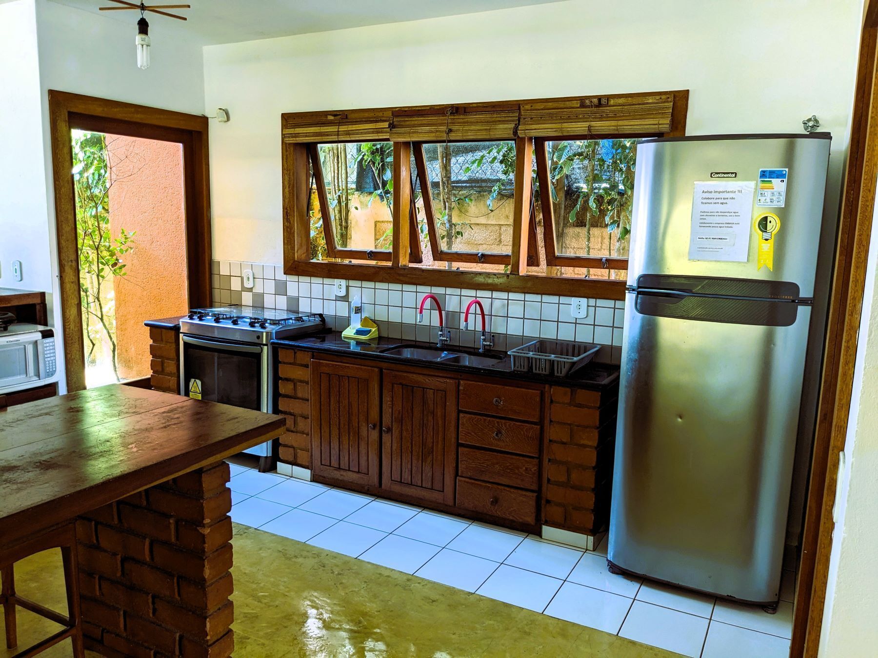A kitchen with a gas stove, sink, wooden cabinets, a refrigerator, and a window overlooking greenery.