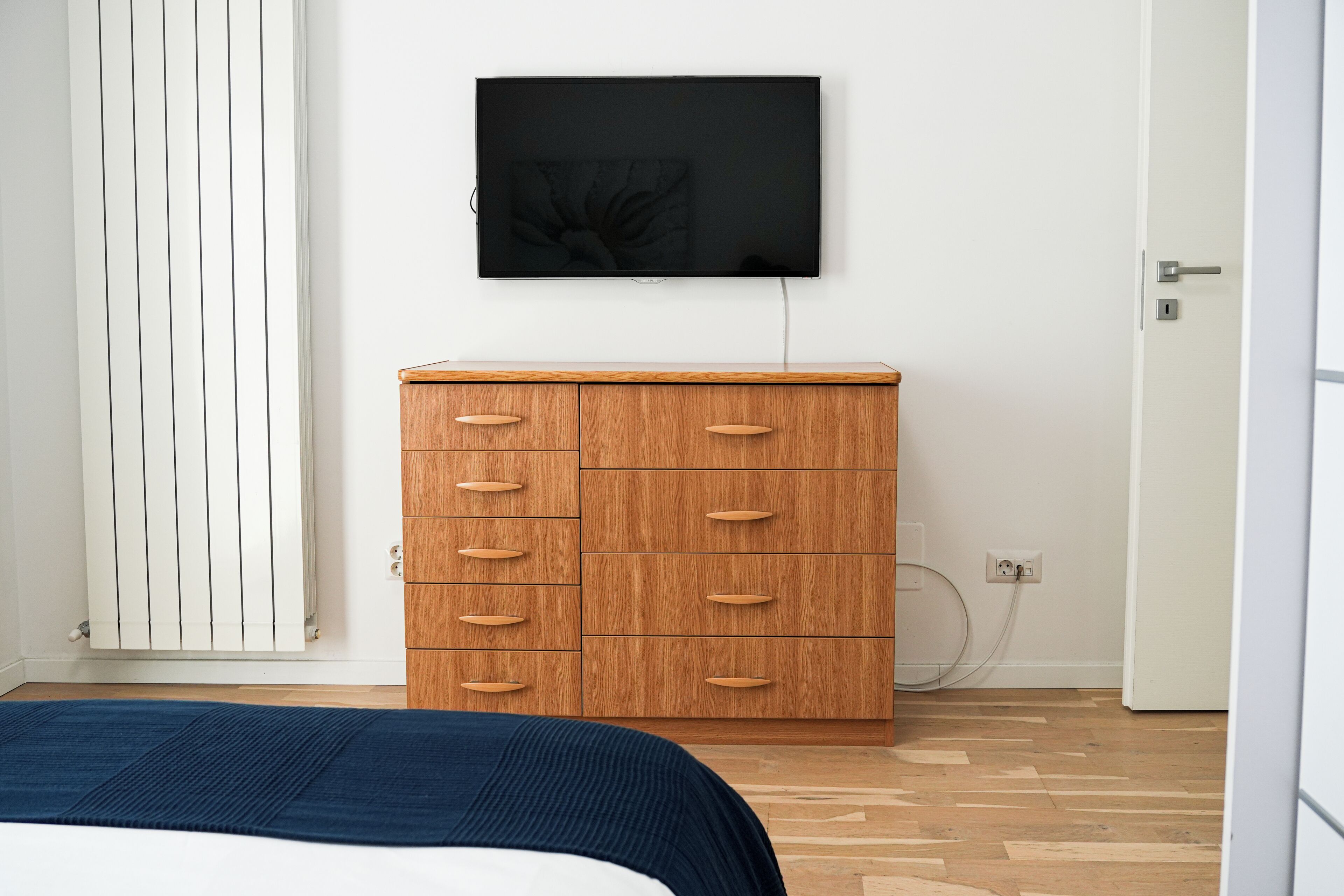Een slaapkamer met een bed, een houten dressoir, een aan de muur bevestigde televisie en een radiator.