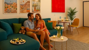 Una pareja en un sofá color turquesa con un plato de aperitivos, un cuadro rojo y una mesita con velas.