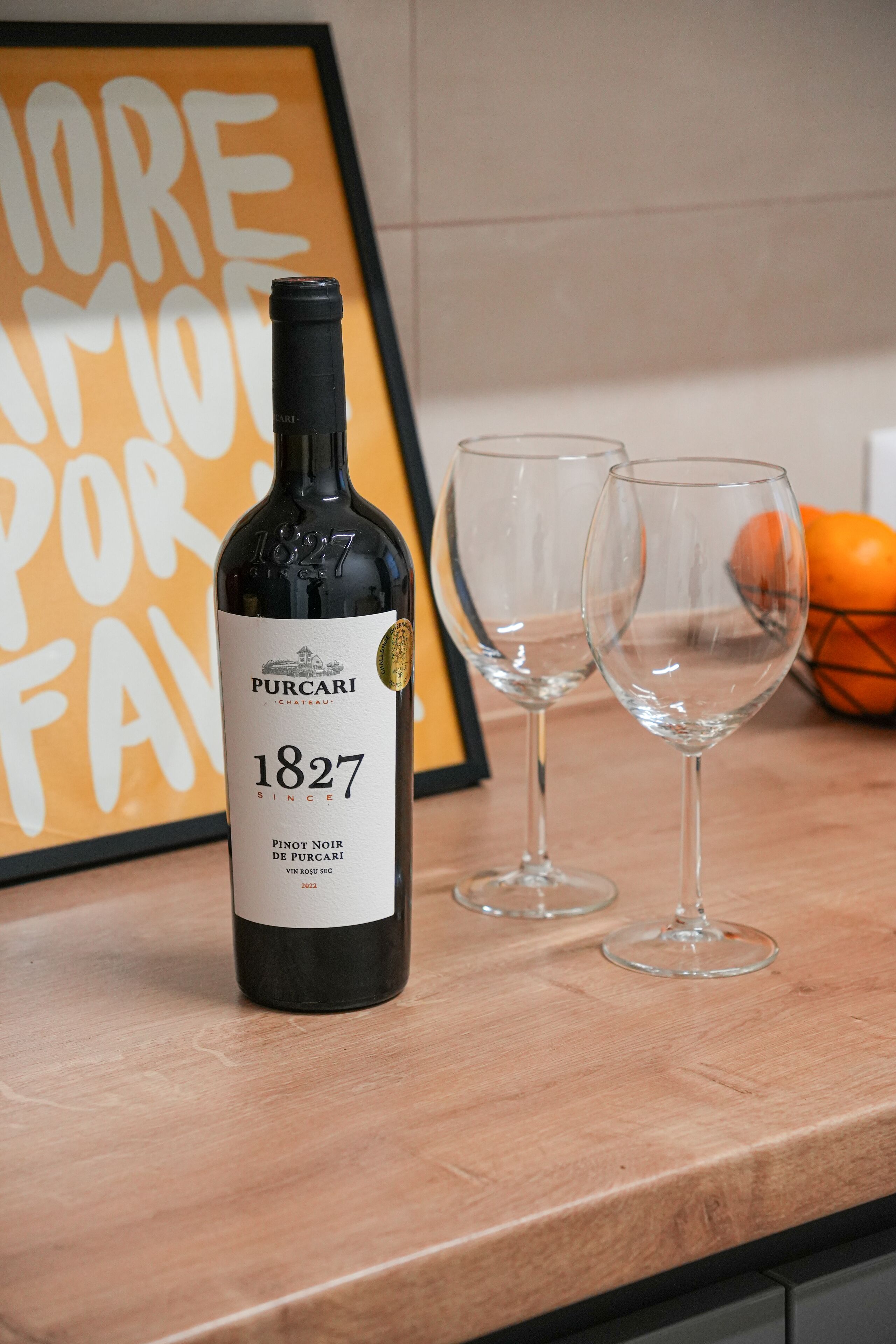 Une bouteille de vin Pinot Noir de Pucari, millésime 1827, est posée sur un comptoir de cuisine, à côté de deux verres à vin vides. Un panneau encadré est visible en arrière-plan.