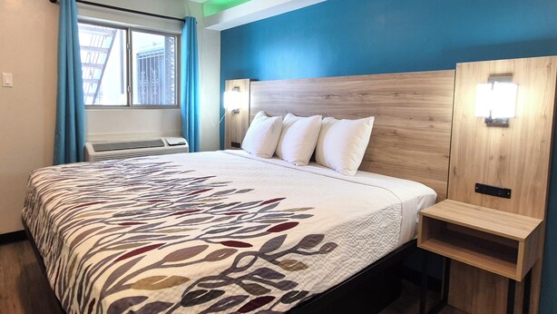 A hotel room with a bed, a window with curtains, a wall-mounted light, and a wooden nightstand. - Exclusivo Inn by OYO Grand Prairie, TX (Grand Prairie)