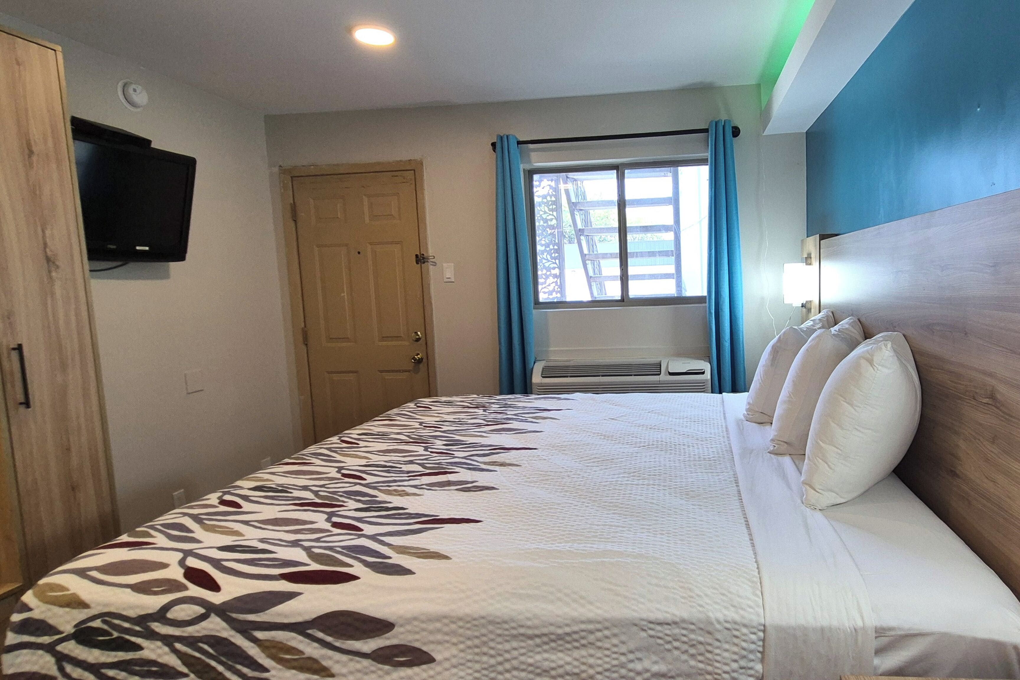 A hotel room with a bed, a television mounted on the wall, a window with curtains, and an air conditioning unit.