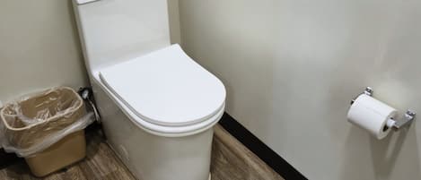 Des toilettes avec un abattant blanc, une poubelle en plastique et un porte-rouleau de papier toilette fixé au mur.