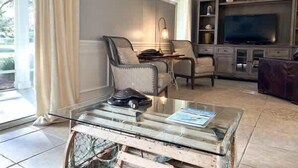 A living room with a glass coffee table, two armchairs, a TV, and a bookshelf.