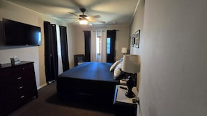 A bedroom with a bed, a flat-screen TV, a ceiling fan, a bedside lamp, and a dresser.