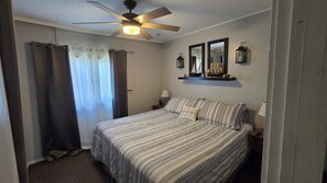 A bedroom with a bed, a ceiling fan, a nightstand, a lamp, a mirror, and a shelf with decorative items.