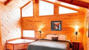 A cozy cabin room with a wooden interior, a large bed, a built-in bathtub, and two wall-mounted lamps. - Secluded Escape | Spacious Deck (Ruidoso)