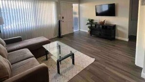 A living room with a sofa, a glass coffee table, and a television on the wall. - Modern, Comfortable 3bd w/ Backyard Space & Grill (Torrance)