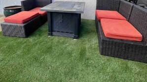 An outdoor patio area with wicker furniture and a table, surrounded by artificial grass. - Modern, Comfortable 3bd w/ Backyard Space & Grill (Torrance)