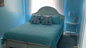 Un dormitorio con cama, una ventana con persianas, una mesita de noche con lámpara y una puerta.