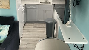 Cocina compacta con armarios grises, un lavamanos y una encimera. Hay un cubo de basura negro y una mesita con una silla.