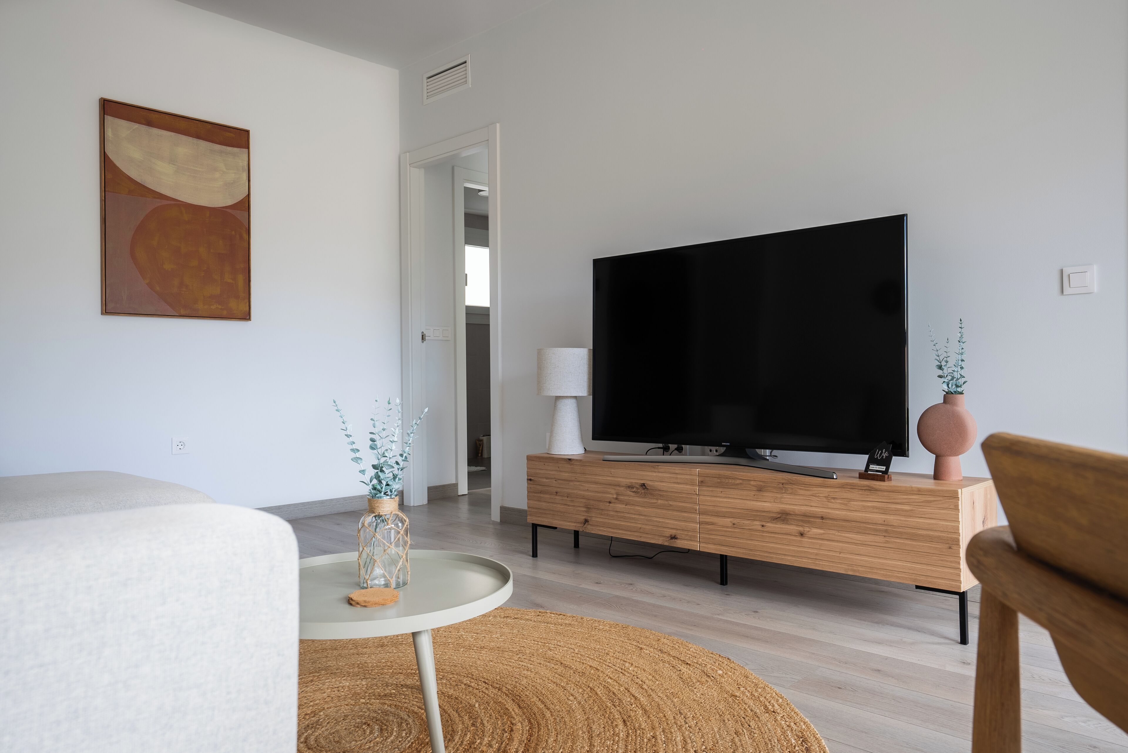 Een moderne woonkamer met een flatscreen televisie op een houten kast, een ronde salontafel met een vaas, en een groot abstract schilderij aan de muur.