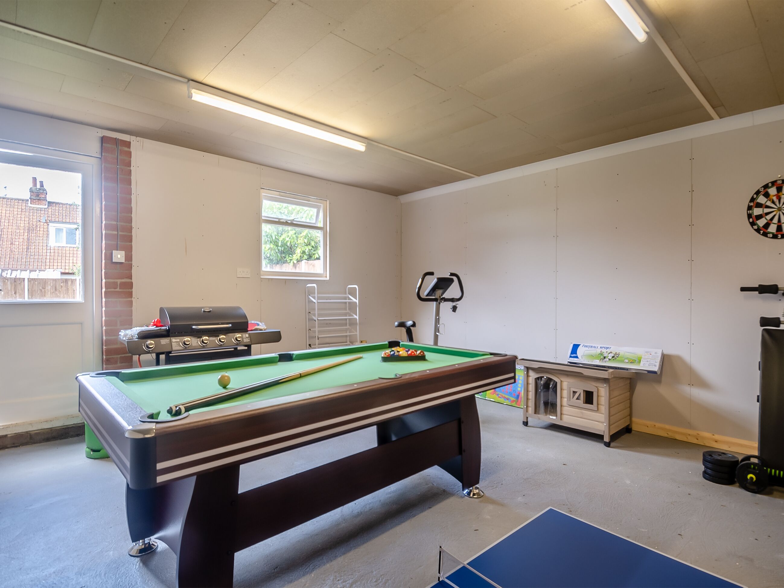 A room with a pool table, a dartboard, a treadmill, and a barbecue grill.