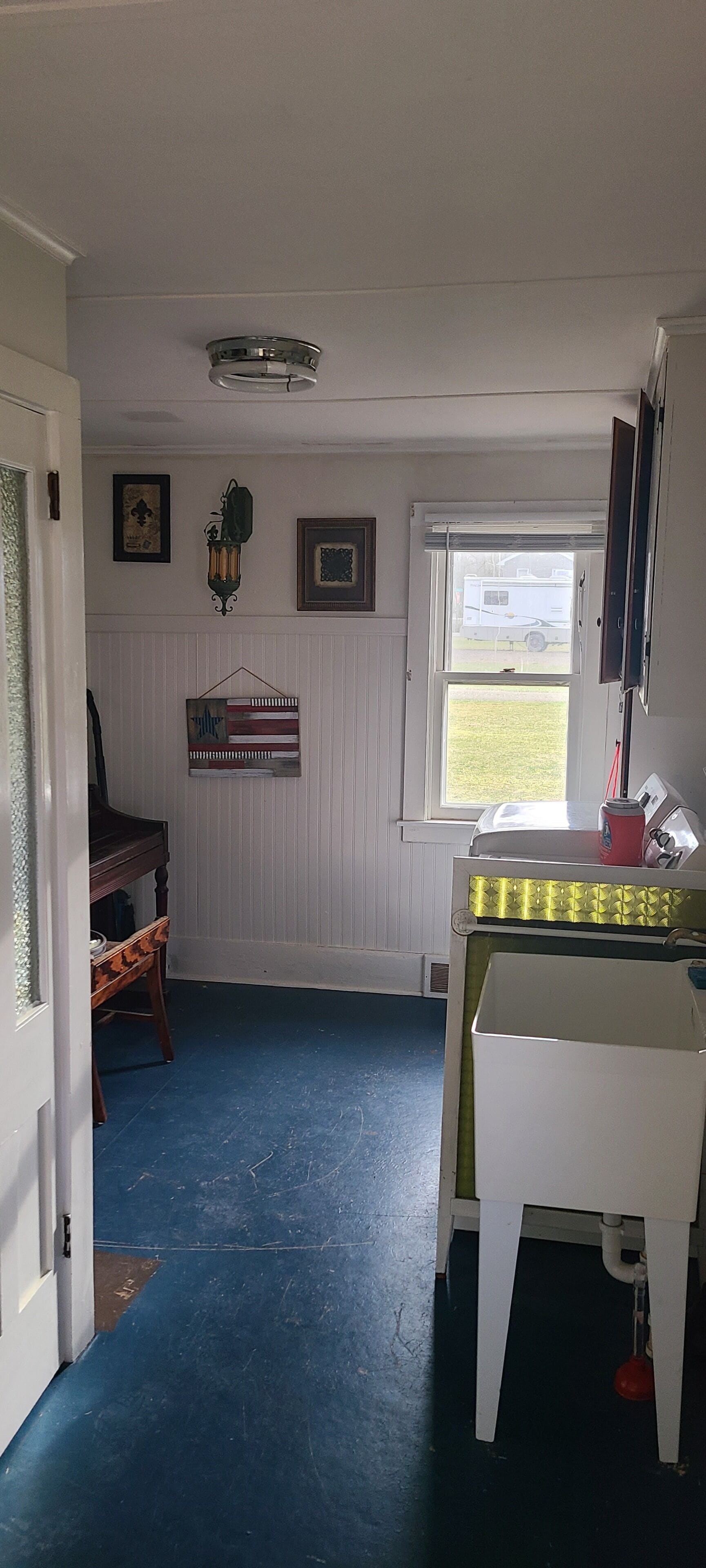 A room with a blue floor, a piano, a white table with a yellow tablecloth, a window with a view of a trailer, and wall-mounted decorations.