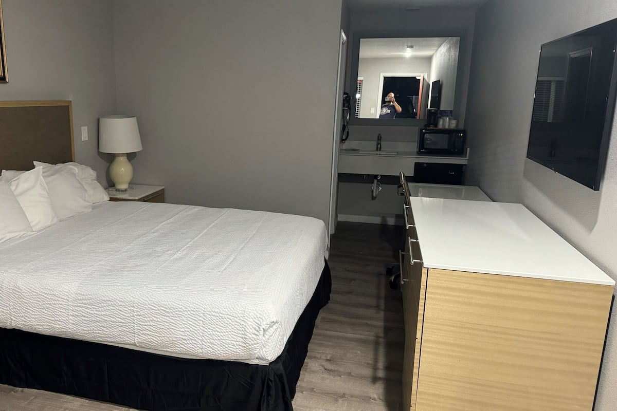 A hotel room with a bed, a nightstand, a lamp, a mirror, a TV, and a kitchenette.