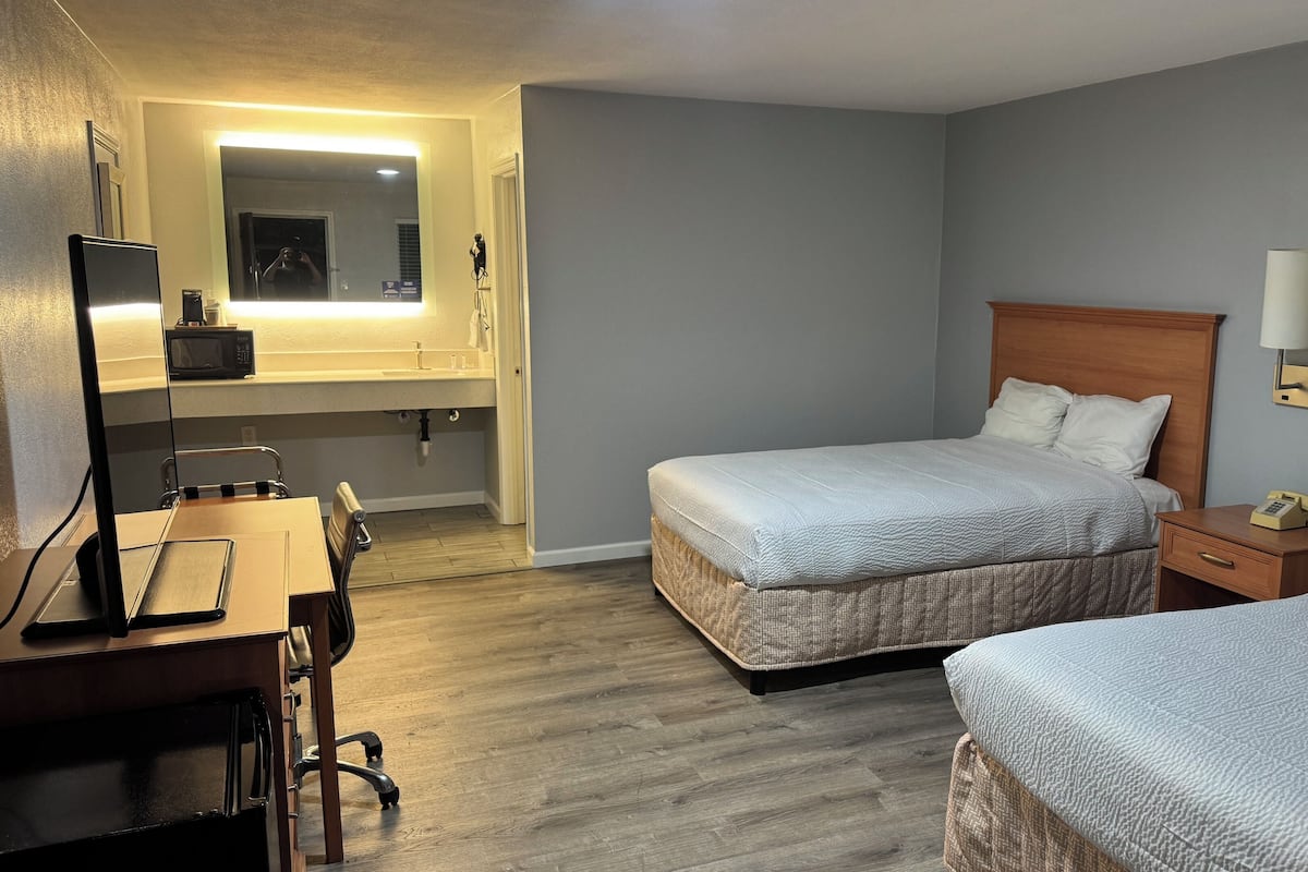 A hotel room with two beds, a desk with a chair, a mirror, and a bedside table with a lamp.