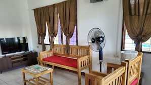 Ein Zimmer mit einer Holz-Couch, einem passenden Sessel, einem kleinen Tisch, einem Fernseher und einem Standventilator.