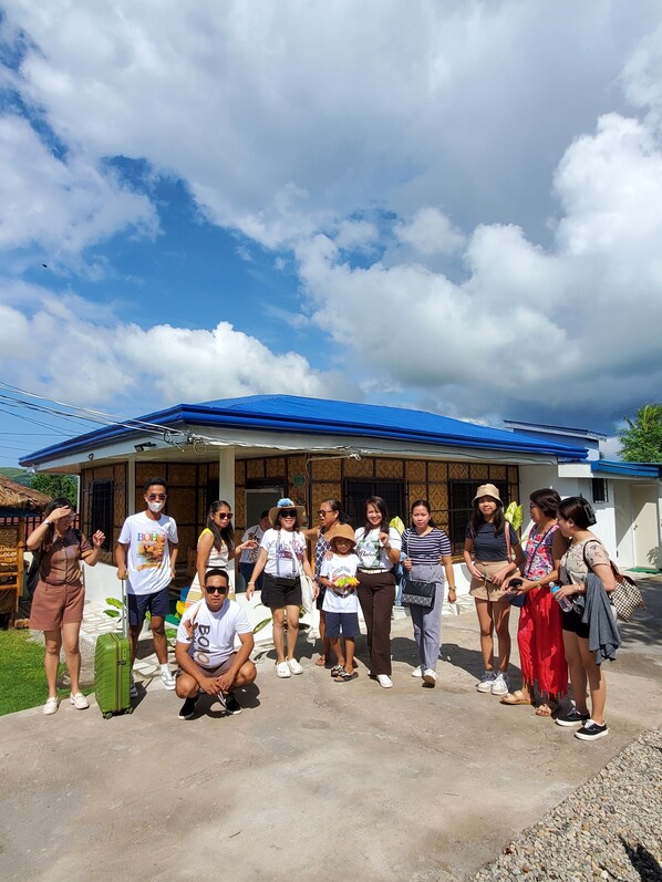 A group of people standing in front of a building with a blue roof and a wooden facade. - Sleeps up to 8 , additional 6 dollars fee over 8 bungalow house. (Tagbilaran)