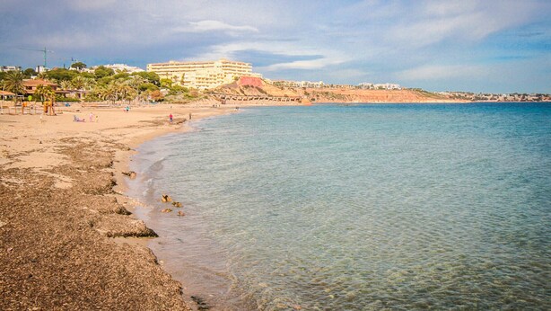 Une plage avec une eau claire, un rivage sablonneux et un grand bâtiment en arrière-plan.