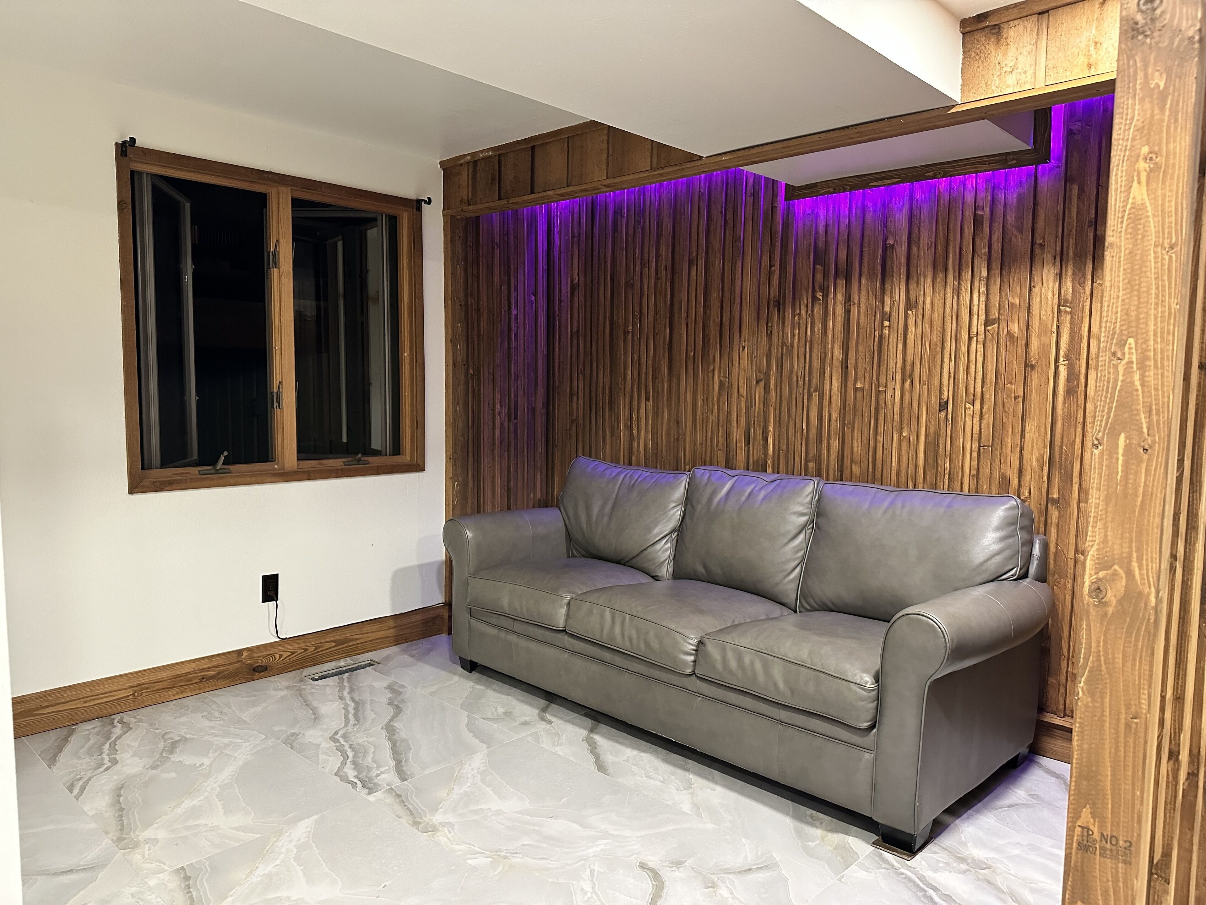 A room with a grey leather sofa, wooden panel wall, and a window with blinds.