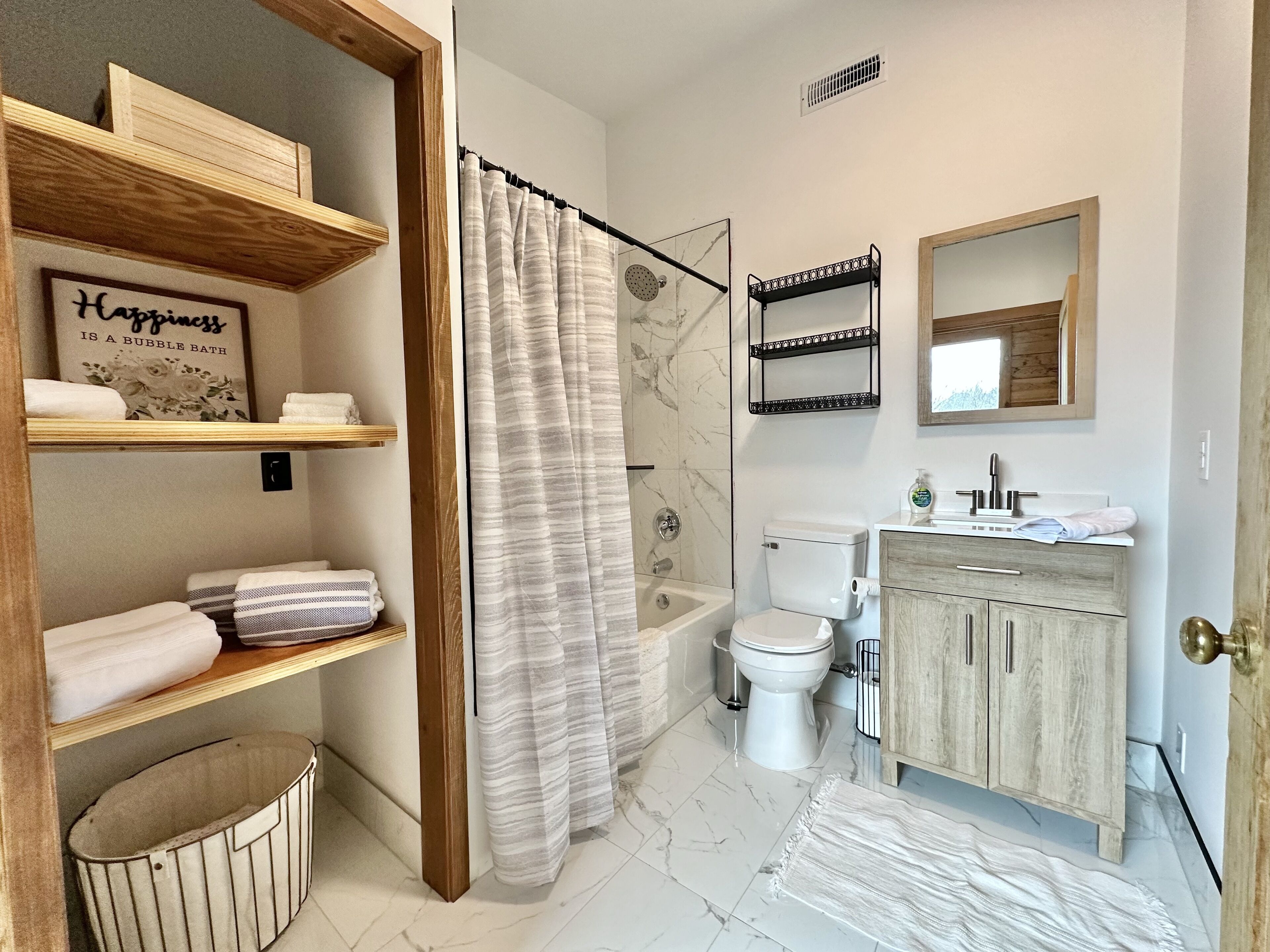 Ein Badezimmer mit Badewanne, Toilette und Waschbecken. Es gibt Regale mit Handtüchern und ein gerahmtes Zitat an der Wand.