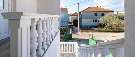 A residential building with a white balcony railing, a staircase leading to the entrance, and a view of neighboring houses.