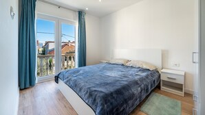 Un dormitorio con cama, mesita de noche, ventana con cortinas y vista a edificios.