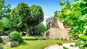 A stone building with a sloped roof, surrounded by lush greenery and a gravel path. - La Metairie De La Garenne (Nadaillac De Rouge)