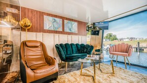Una sala de estar moderna con un sillón de cuero, un sofá de terciopelo y una butaca rosa. Hay una mesa de centro de vidrio y un ventanal con vistas a un cuerpo de agua.