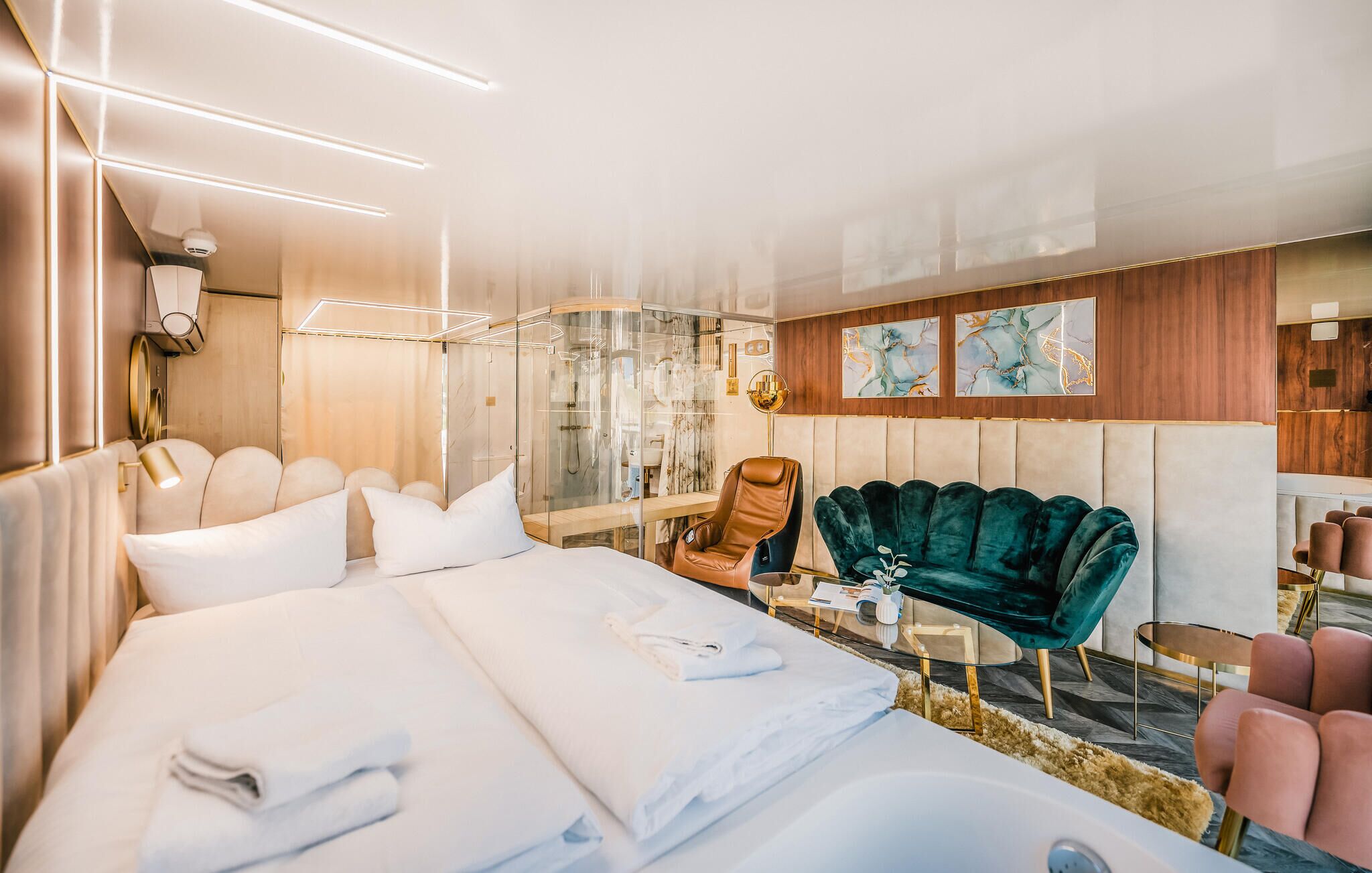 Una habitación de hotel moderna con una cama grande, una zona de estar con un sillón de terciopelo verde, un box de ducha de vidrio y obras de arte en las paredes.