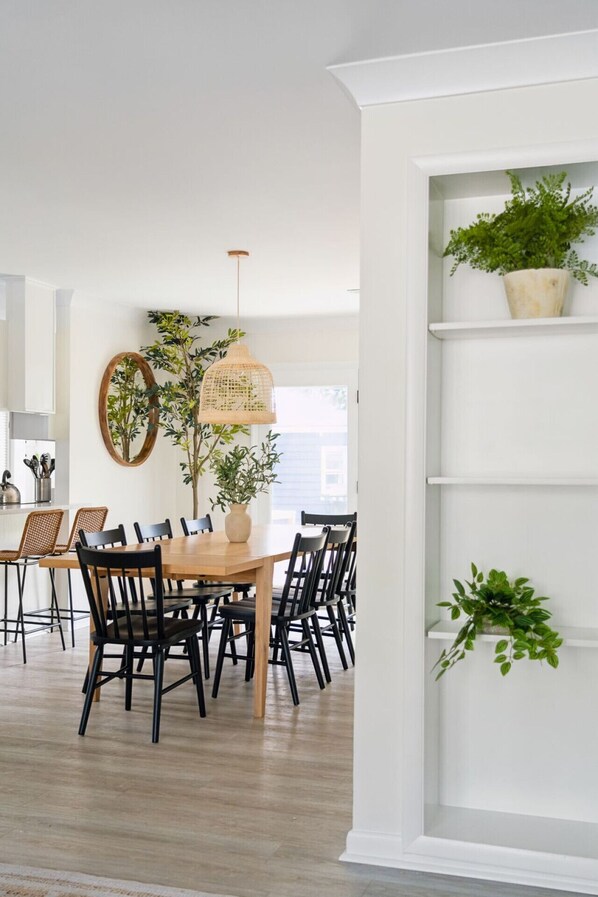 A dining area with a wooden table and black chairs, surrounded by white walls and a white shelving unit with plants.