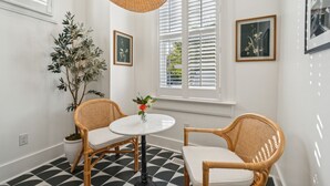 Een kamer met een zwart-wit geruite vloer, twee rieten stoelen, een ronde tafel, een kamerplant en twee ingelijste foto’s aan de muur.