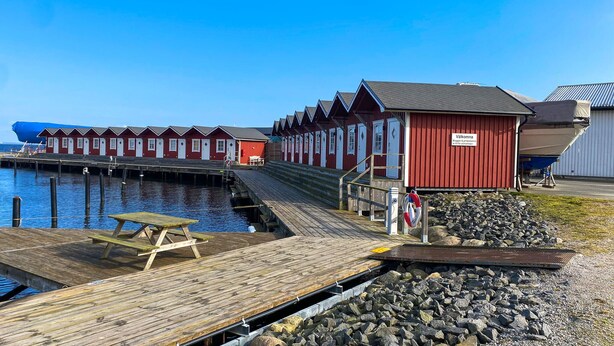 Une rangée de bâtiments en bois peints en rouge, avec un quai s’avançant dans une eau calme, une table de pique-nique et un bouée de sauvetage.