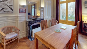 A dining area with a wooden table and chairs, a marble fireplace, and a large window with curtains. - ⚓ L’Avant Port | Stunning View of Honfleur’s Outer Harbor | 65 m² | 4 Guest (Honfleur)