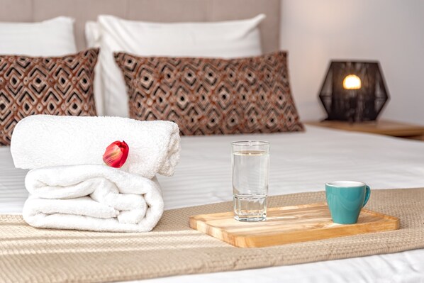 A neatly made bed with patterned pillows, a folded white towel with a red flower, a glass of water, and a teal mug on a wooden tray.