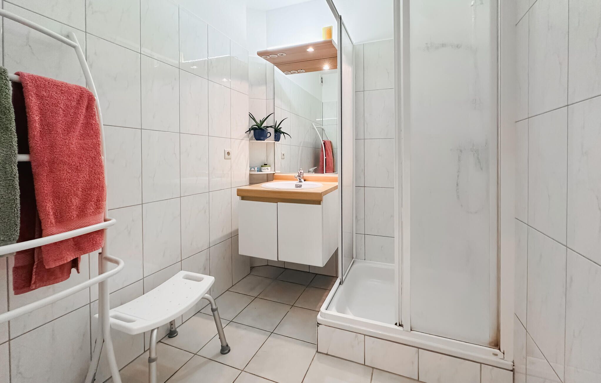 Um banheiro moderno com azulejos brancos, box de chuveiro, pia com bancada de madeira e um pequeno banco branco.