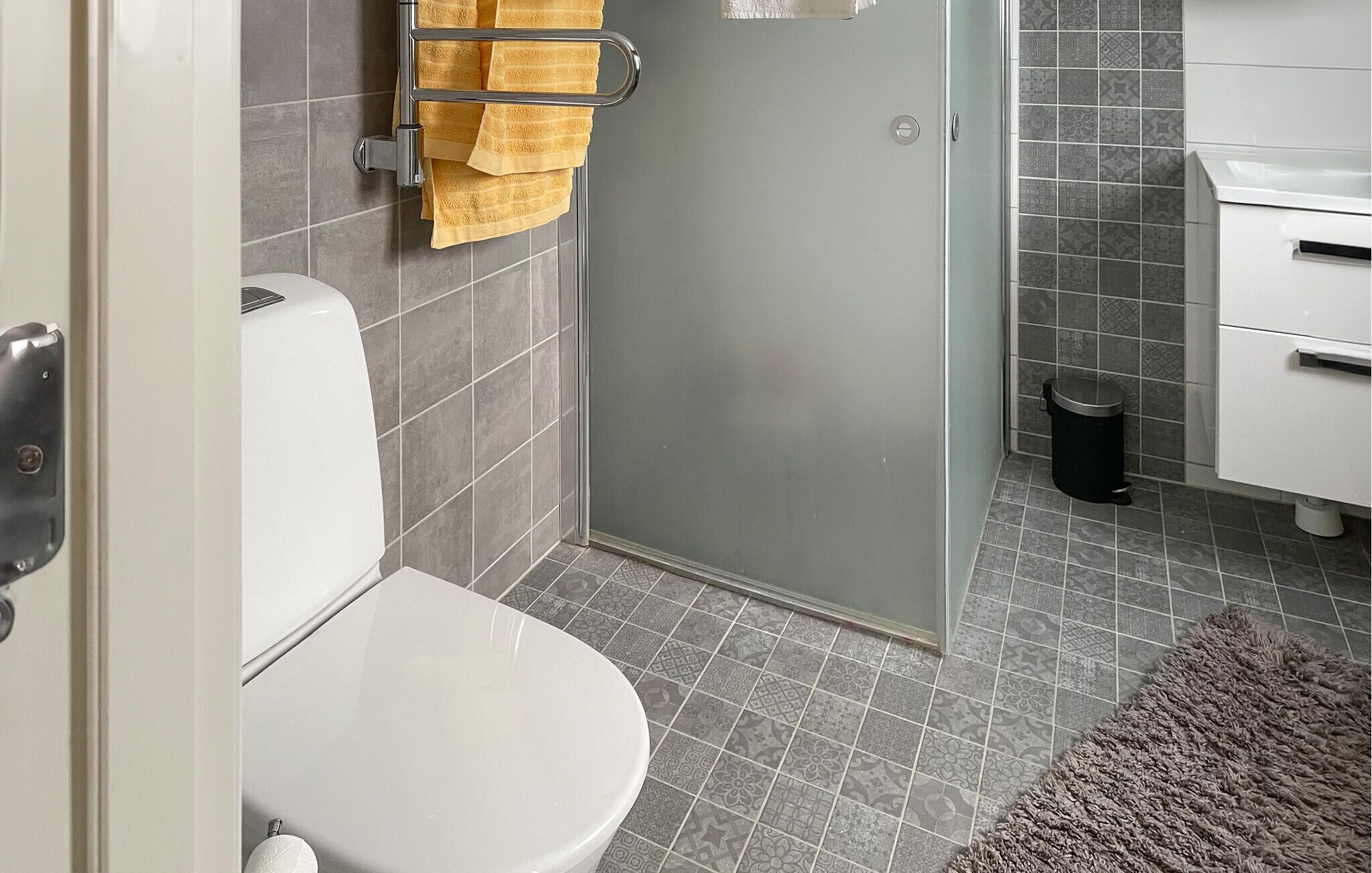 A bathroom with a toilet, a glass shower enclosure, a towel rack with yellow towels, a trash bin, and a patterned rug.