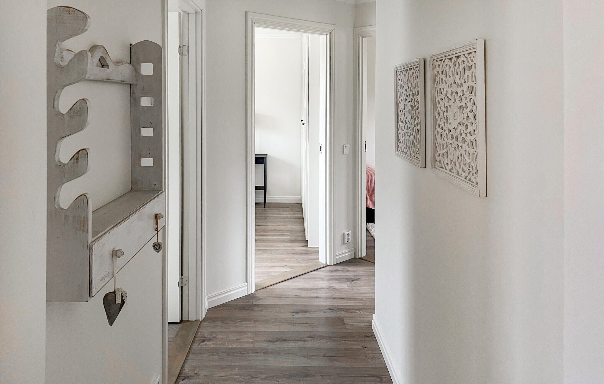 A hallway with a wooden floor, white walls, and a door leading to another room. There are two decorative wall panels on the right.