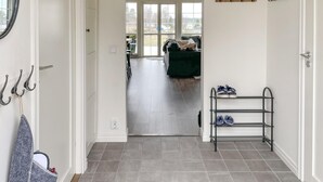 Un couloir doté d’un sol carrelé de couleur grise, d’un porte-chaussures en métal rempli de chaussures et d’une porte donnant sur une pièce où se trouvent un canapé vert foncé et une fenêtre.